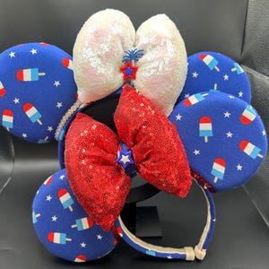 K&ouml;nnte beinhalten: Ein festliches Stirnband mit blauen, roten und wei&szlig;en Elementen. Das Design umfasst blaue kreisf&ouml;rmige Ohren mit Eis am Stiel-Grafiken und wei&szlig;en Sternen. Eine rote Pailletten-Schleife und wei&szlig;e Pailletten-Ohren vervollst&auml;ndigen das patriotische Thema.