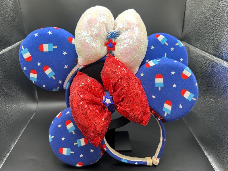 K&ouml;nnte beinhalten: Ein festliches Stirnband mit blauen, roten und wei&szlig;en Elementen. Das Design umfasst blaue kreisf&ouml;rmige Ohren mit Eis am Stiel-Grafiken und wei&szlig;en Sternen. Eine rote Pailletten-Schleife und wei&szlig;e Pailletten-Ohren vervollst&auml;ndigen das patriotische Thema.