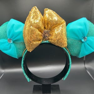 May include: A teal and gold sequined headband with large mouse ears. The ears are teal with a bow and a jewel in the centre. The centre bow is gold with sequins and a small brown embellishment.