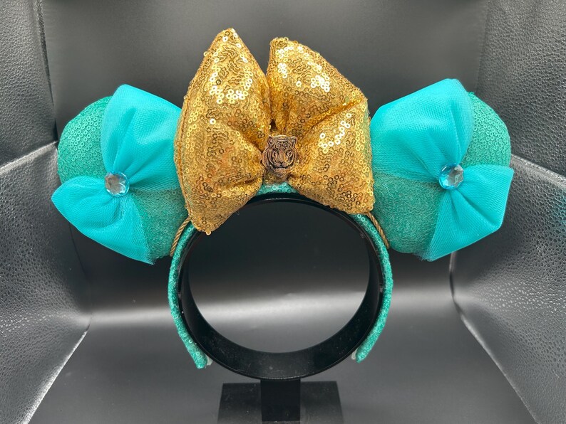 May include: A teal and gold sequined headband with large mouse ears. The ears are teal with a bow and a jewel in the centre. The centre bow is gold with sequins and a small brown embellishment.