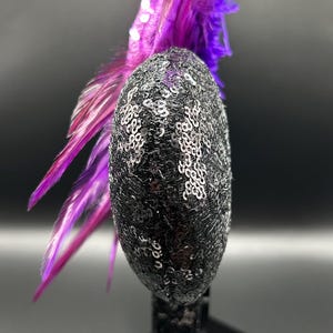 May include: A headband featuring a circular ear-shaped component covered in black sequins. Attached to the side of the ear is a cluster of vibrant purple and magenta feathers that extend upwards and outwards from the top of the headband structure.
