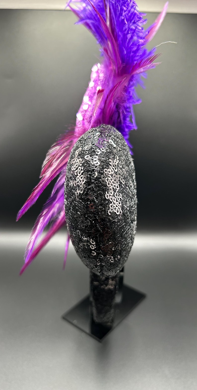 May include: A headband featuring a circular ear-shaped component covered in black sequins. Attached to the side of the ear is a cluster of vibrant purple and magenta feathers that extend upwards and outwards from the top of the headband structure.