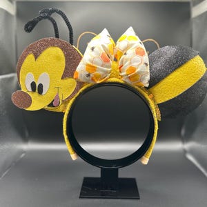 May include: A pair of yellow and black bee-themed Minnie Mouse ears with a brown and yellow bee face on one ear and a black and yellow striped bee on the other ear. The ears have a honeycomb patterned bow.