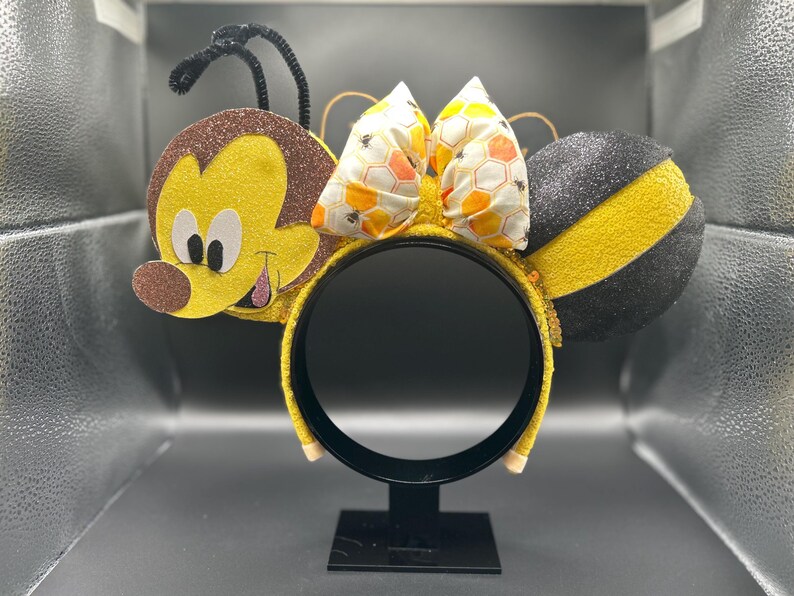 May include: A pair of yellow and black bee-themed Minnie Mouse ears with a brown and yellow bee face on one ear and a black and yellow striped bee on the other ear. The ears have a honeycomb patterned bow.