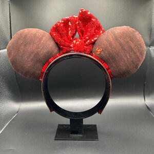 May include: A headband featuring two round brown ears with a textured surface. A large, bright red sequined bow sits between the ears. The headband frame is covered in red sequins, and a small orange gem shaped like a mouse head is attached to the right ear.