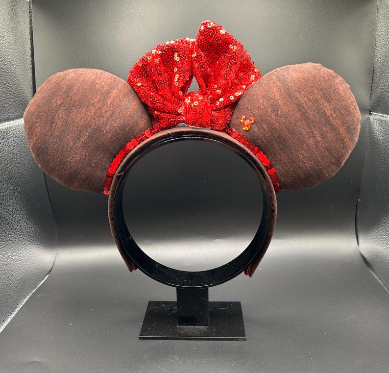 May include: A headband featuring two round brown ears with a textured surface. A large, bright red sequined bow sits between the ears. The headband frame is covered in red sequins, and a small orange gem shaped like a mouse head is attached to the right ear.