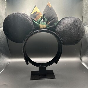 May include: Black mouse ear headband with a camouflage bow. The ears are round and made of a soft, velvety material. The bow features a green, brown, and tan camouflage pattern. The headband is black and has a sturdy construction.