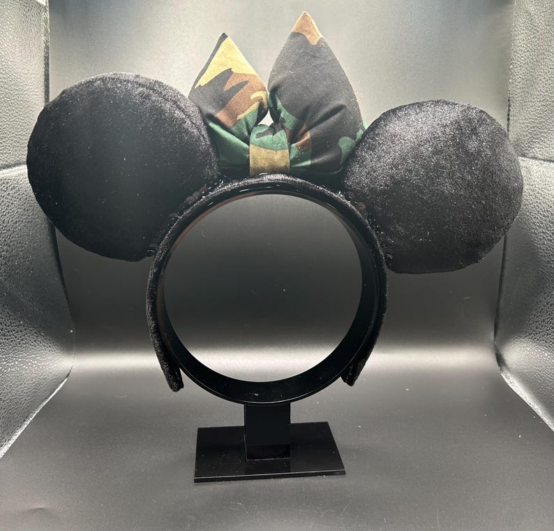 May include: Black mouse ear headband with a camouflage bow. The ears are round and made of a soft, velvety material. The bow features a green, brown, and tan camouflage pattern. The headband is black and has a sturdy construction.