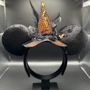 Puede incluir: Una diadema con dos grandes orejas de terciopelo negro y un sombrero de bruja central. El sombrero est&aacute; cubierto de encaje naranja con plumas negras y una banda de pedrer&iacute;a plateada. Un peque&ntilde;o icono naranja de tres c&iacute;rculos est&aacute; en la oreja.