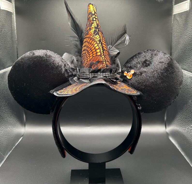 Puede incluir: Una diadema con dos grandes orejas de terciopelo negro y un sombrero de bruja central. El sombrero est&aacute; cubierto de encaje naranja con plumas negras y una banda de pedrer&iacute;a plateada. Un peque&ntilde;o icono naranja de tres c&iacute;rculos est&aacute; en la oreja.