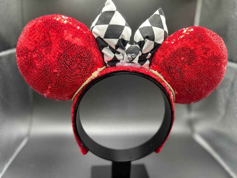 May include: Red sequined Minnie Mouse ears with a black and white checkered bow. The bow has a small black and white character in the center.