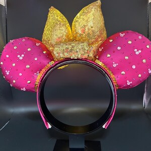 May include: A pair of hot pink mouse ears with a gold sequined bow and trim. The ears are embellished with small, sparkling rhinestones. The headband is black with a pink trim.