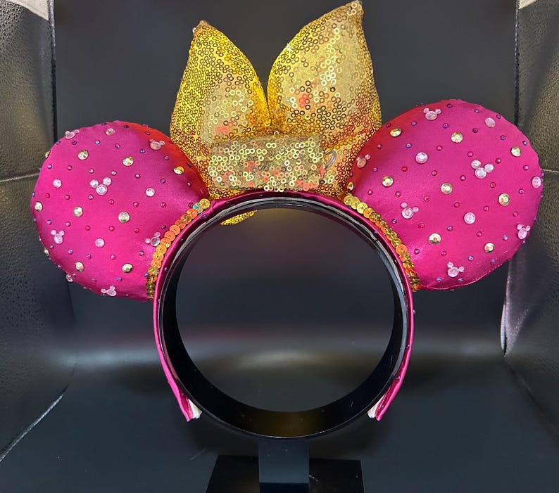 May include: A pair of hot pink mouse ears with a gold sequined bow and trim. The ears are embellished with small, sparkling rhinestones. The headband is black with a pink trim.