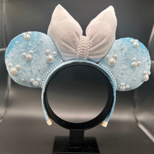 May include: Light blue velvet mouse ear headband with pearl accents. The ears are decorated with pearl embellishments and Mickey Mouse-shaped pearl clusters. A large, light gray velvet bow is centered on the headband, also adorned with pearls.