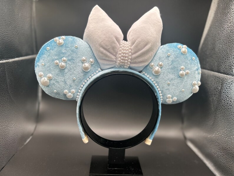 May include: Light blue velvet mouse ear headband with pearl accents. The ears are decorated with pearl embellishments and Mickey Mouse-shaped pearl clusters. A large, light gray velvet bow is centered on the headband, also adorned with pearls.