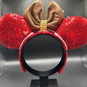 May include: A headband featuring two large, round ears covered in bright red sequins. A brown fabric bow sits between the ears, accented with a small gold sequin centre. A tiny gold-toned Mickey Mouse icon is attached to the left red ear.