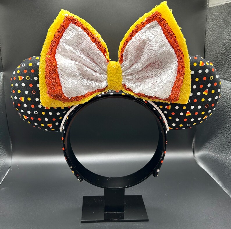 May include: A festive headband with mouse ears. The ears are black with a pattern of candy corn and polka dots. The large bow is white with yellow and orange sequin trim. The headband is black with a white trim.