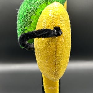 May include: A whimsical headband featuring a black faux fur accent, a vibrant green sequined section, and a yellow sequined section. A black fuzzy wire adds a playful touch. This accessory is perfect for themed events.