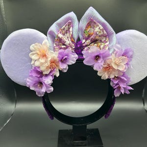 Puede incluir: Una diadema con dos grandes orejas circulares de color morado claro y un lazo central de tela iridiscente con motivos florales. La base de las orejas est&aacute; adornada con un grupo de flores artificiales en tonos rosa claro y morado.