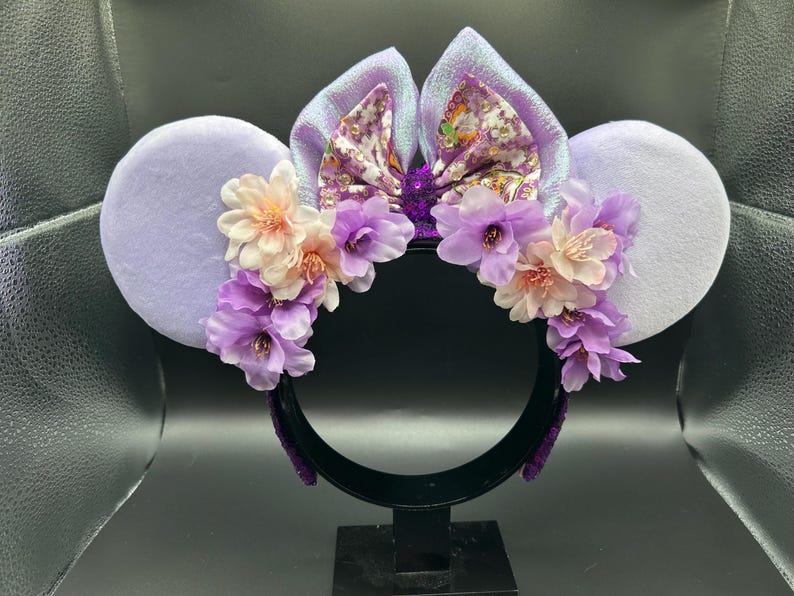 Puede incluir: Una diadema con dos grandes orejas circulares de color morado claro y un lazo central de tela iridiscente con motivos florales. La base de las orejas est&aacute; adornada con un grupo de flores artificiales en tonos rosa claro y morado.