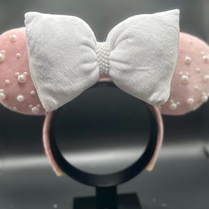 May include: Pink Minnie Mouse ears with a white velvet bow and pearl embellishments.