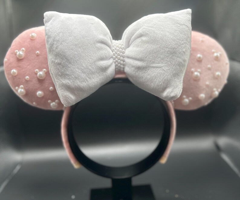 May include: Pink Minnie Mouse ears with a white velvet bow and pearl embellishments.
