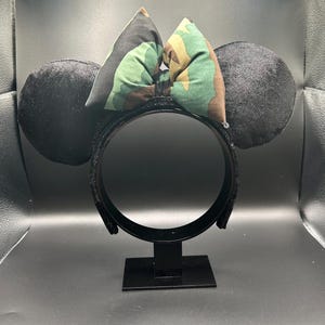 May include: Black mouse ear headband with black velvet ears and a camouflage bow. The headband is on a black stand. The bow features green, brown, and tan camouflage fabric. The ears are round and plush.