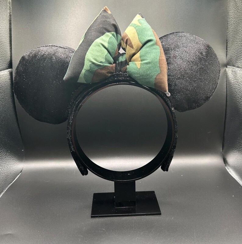 May include: Black mouse ear headband with black velvet ears and a camouflage bow. The headband is on a black stand. The bow features green, brown, and tan camouflage fabric. The ears are round and plush.