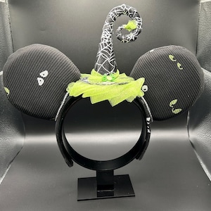 May include: Black Halloween-themed headband with large, round ears. The ears have white eye designs. A witch hat with a spiderweb design and lime green accents sits atop the headband. The headband is displayed on a black stand.