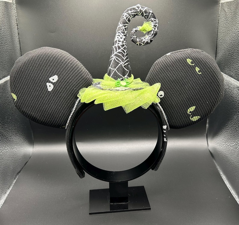 May include: Black Halloween-themed headband with large, round ears. The ears have white eye designs. A witch hat with a spiderweb design and lime green accents sits atop the headband. The headband is displayed on a black stand.