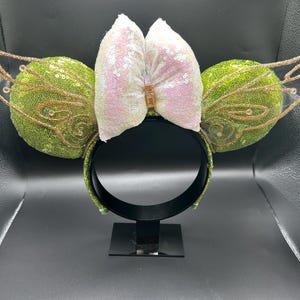 Op de afbeelding: Een haarband met twee ronde oren bedekt met groene lovertjes, aan weerszijden geaccentueerd met gouden glitter fee&euml;nvleugels. Een grote, iriserende witte lovertjesstrik zit in het midden, vastgehouden door een klein goudkleurig rechthoekig stukje.