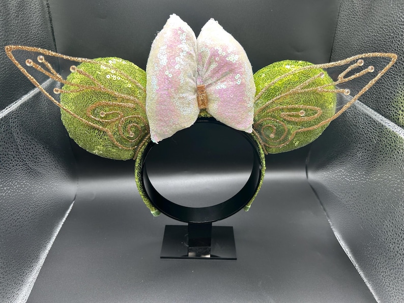 Op de afbeelding: Een haarband met twee ronde oren bedekt met groene lovertjes, aan weerszijden geaccentueerd met gouden glitter fee&euml;nvleugels. Een grote, iriserende witte lovertjesstrik zit in het midden, vastgehouden door een klein goudkleurig rechthoekig stukje.