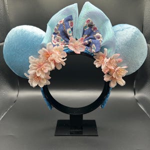 Peut inclure: Un serre-t&ecirc;te avec deux grandes oreilles en velours bleu clair, un n&oelig;ud central bleu iris&eacute; et un groupe de fleurs de cerisier artificielles rose p&acirc;le dispos&eacute;es sur le devant. La base du serre-t&ecirc;te est noire et recouverte de sequins bleus.