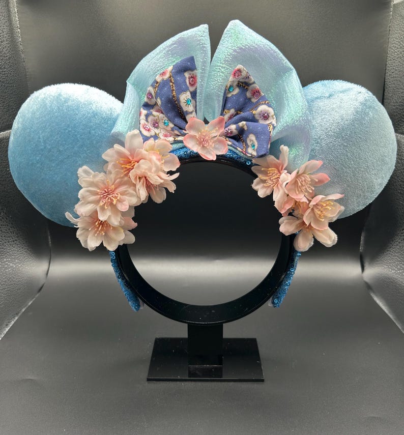 Peut inclure: Un serre-t&ecirc;te avec deux grandes oreilles en velours bleu clair, un n&oelig;ud central bleu iris&eacute; et un groupe de fleurs de cerisier artificielles rose p&acirc;le dispos&eacute;es sur le devant. La base du serre-t&ecirc;te est noire et recouverte de sequins bleus.