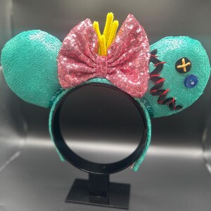 May include: Teal sequined mouse ears with a pink sequined bow and yellow accents. One ear features black stitching, a yellow cross button, and a blue button. The headband is black.