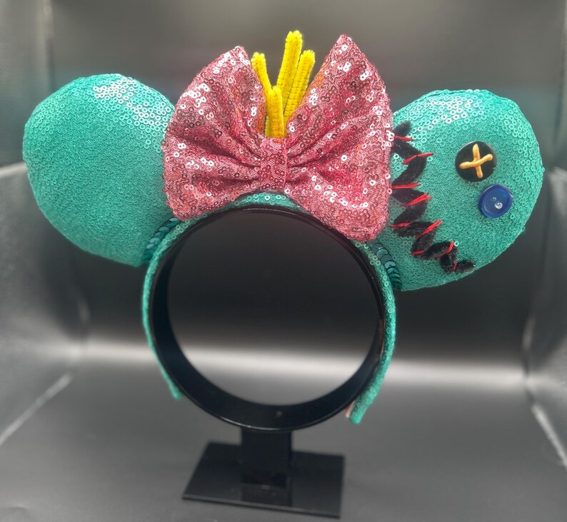 May include: Teal sequined mouse ears with a pink sequined bow and yellow accents. One ear features black stitching, a yellow cross button, and a blue button. The headband is black.