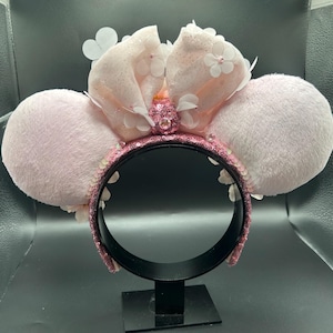 Op de afbeelding: Een haarband met twee grote, pluizige, lichtroze ronde oren. Een centrale, extra grote, glitterende lichtroze stoffen strik is versierd met kleine witte bloemapplicaties en een centrale roze strass-cluster. Het frame is bedekt met roze lovertjes.