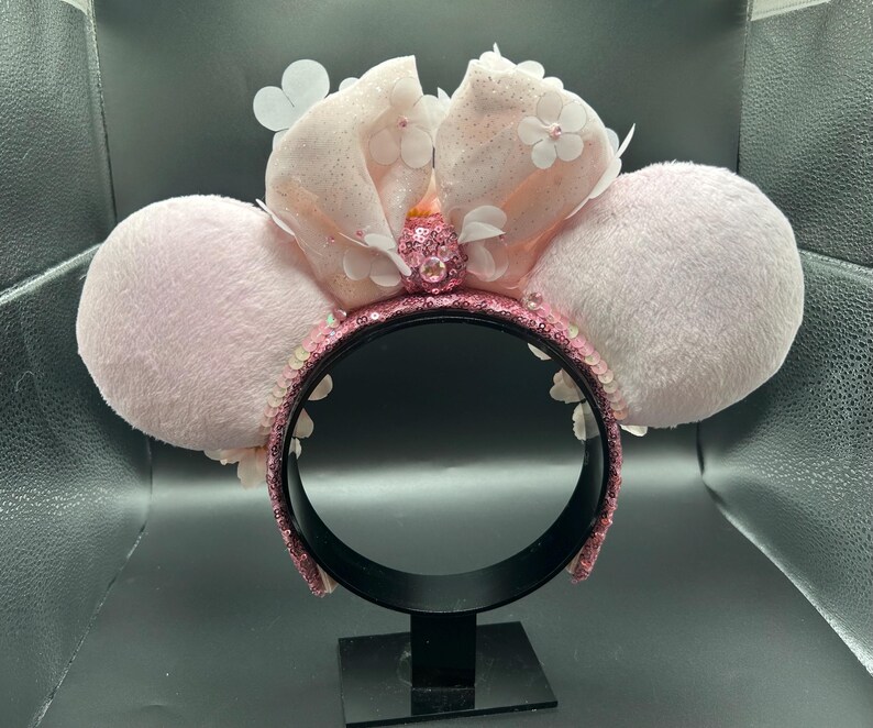 Op de afbeelding: Een haarband met twee grote, pluizige, lichtroze ronde oren. Een centrale, extra grote, glitterende lichtroze stoffen strik is versierd met kleine witte bloemapplicaties en een centrale roze strass-cluster. Het frame is bedekt met roze lovertjes.
