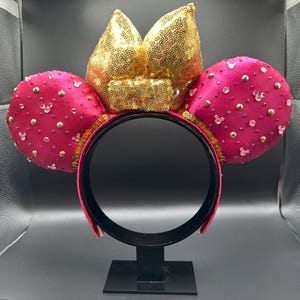 May include: A magenta-colored headband with two round ears and a bow. The ears are embellished with small, sparkling accents and the bow is covered in gold sequins. The headband is black and the ears are decorated with small, clear Mickey Mouse silhouettes.