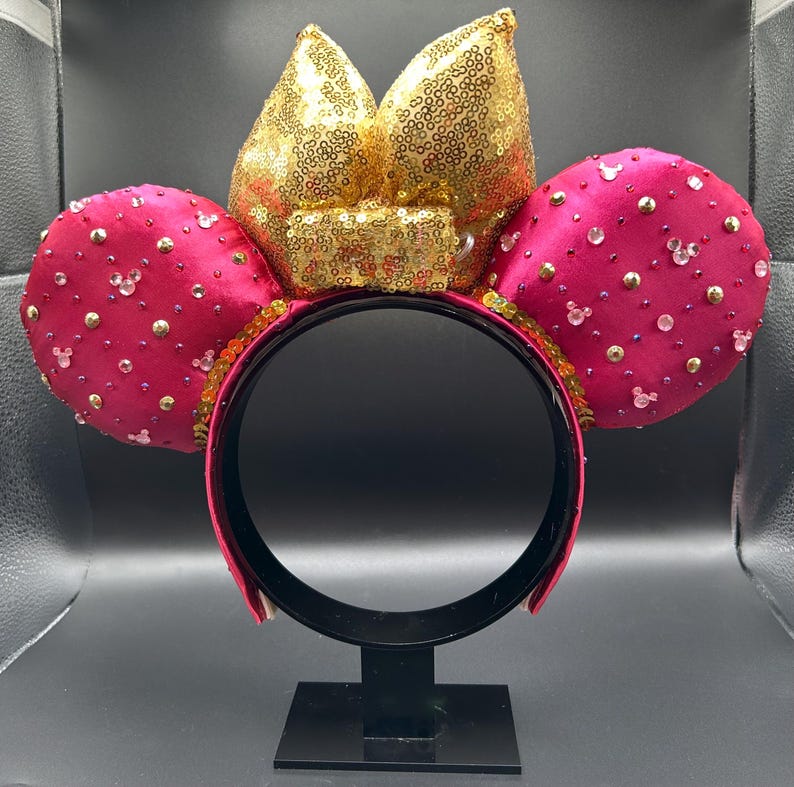 May include: A magenta-colored headband with two round ears and a bow. The ears are embellished with small, sparkling accents and the bow is covered in gold sequins. The headband is black and the ears are decorated with small, clear Mickey Mouse silhouettes.
