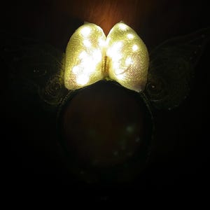 Op de afbeelding: Een haarband met een grote, centrale strik bedekt met glinsterende gouden lovertjes en ge&iuml;ntegreerde witte led-lampjes. De haarband heeft aan weerszijden twee donkere, doorschijnende, vleugelvormige accenten die een lichtgevend effect cre&euml;ren.