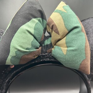 May include: Black headband with two gray plush ears and a camouflage bow. The bow features a green, brown, and black pattern. The headband is decorated with small, shiny sequins.