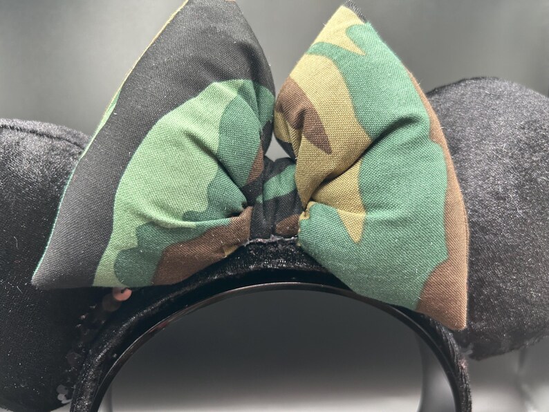 May include: Black headband with two gray plush ears and a camouflage bow. The bow features a green, brown, and black pattern. The headband is decorated with small, shiny sequins.