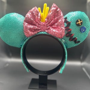 May include: A teal sequined headband with two round ears. A pink sequined bow is centered between the ears, with yellow accents. One ear has a button and stitched details. The headband is on a black stand.