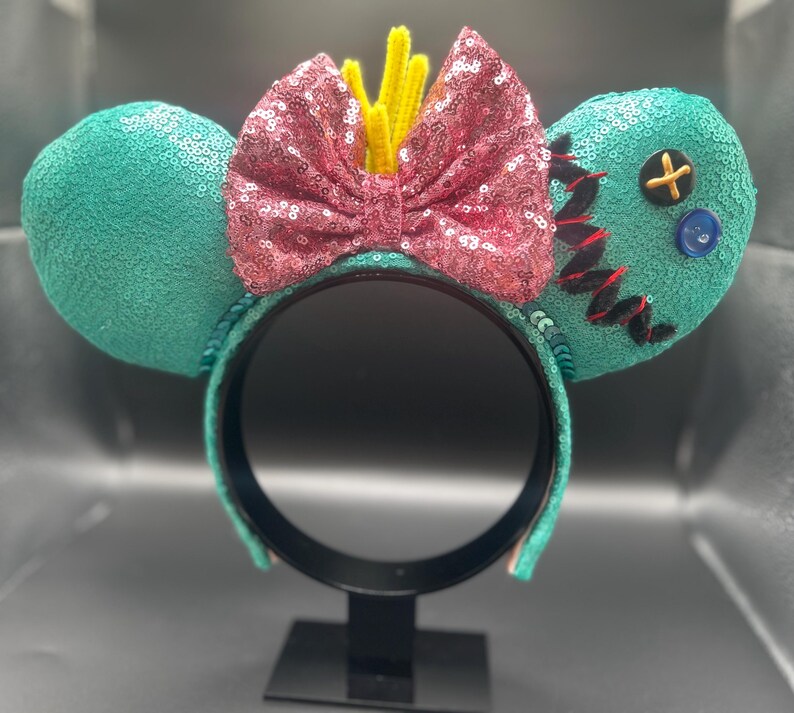 May include: A teal sequined headband with two round ears. A pink sequined bow is centered between the ears, with yellow accents. One ear has a button and stitched details. The headband is on a black stand.