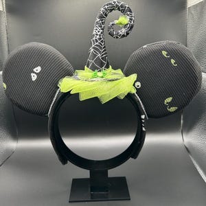 May include: Black Halloween-themed mouse ears headband. The ears are black with white and green accents. A witch hat with a spiderweb design and green tulle is centered on the headband. The headband is on a black stand.