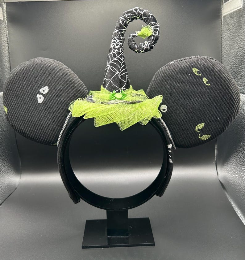 May include: Black Halloween-themed mouse ears headband. The ears are black with white and green accents. A witch hat with a spiderweb design and green tulle is centered on the headband. The headband is on a black stand.