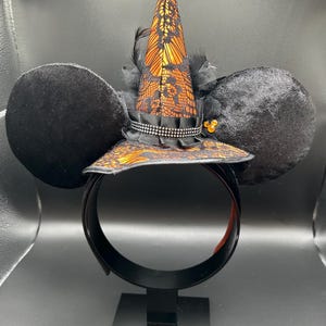 May include: Black mouse ear headband with a witch hat design. The hat is orange and black with lace detailing and a black ribbon. The ears are black and plush. The headband is black with an orange interior.