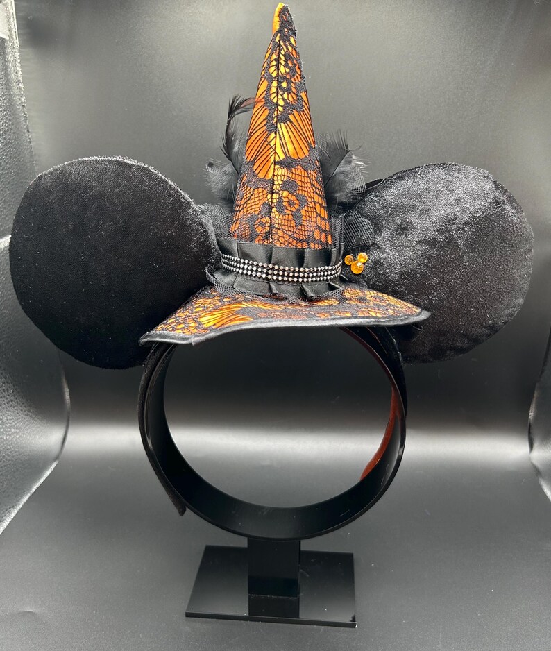 May include: Black mouse ear headband with a witch hat design. The hat is orange and black with lace detailing and a black ribbon. The ears are black and plush. The headband is black with an orange interior.