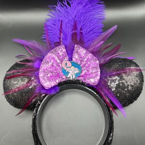 May include: A headband featuring two round ears covered in black sequins, a central light purple sequined bow, and a circular pin depicting a cartoon character. The design is accented with vibrant purple feathers extending upwards and outwards.